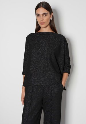 Schwarzes Oberteil mit Dreiviertelärmeln, das aus einem strukturierten Stoff besteht und mit dezenten Glitzerakzenten versehen ist, passend zu maßgeschneiderten schwarzen Hosen.