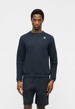 Mand stående frontvendt iført en mørk marineblå sweatshirt med en lille lap på brystet og matchende shorts mod en hvid baggrund.