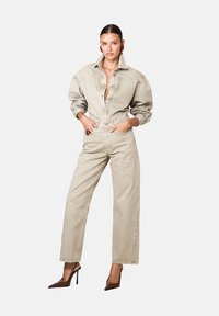 Naine seisab käed taskutes, seljas beež suurus ülikond, sobivad kõrge vöökohaga sirged teksad ja pruunid kontsad.