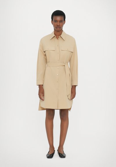 Beige shirt dress with a collar, two chest pockets, button closures, a belted waist, and a rounded hem, styled with black slip-on shoes.