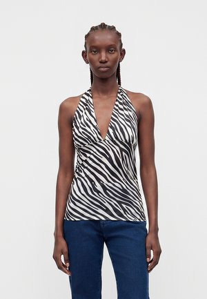 Femme aux cheveux tressés portant un haut dos nu imprimé zèbre noir et blanc et un jean bleu, debout devant un fond uni.