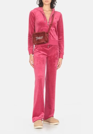 Roze velours trainingspak met een rits hoodie en wijde broekspijpen, gecombineerd met een rode tas met "Juicy" logo en beige instap schoenen.