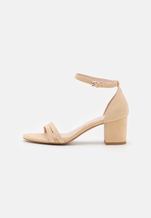Beżowy zamszowy sandał na obcasie z odkrytymi palcami, stabilnym obcasem, paskiem wokół kostki i dwoma paskami z przodu. Gładka faktura i minimalistyczny design.