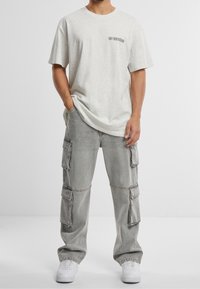 Maglietta in cotone grigio chiaro con scollo rotondo e piccolo logo nero sul petto, abbinata a pantaloni cargo grigi con multiple tasche.