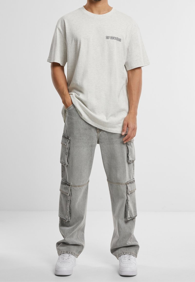 Maglietta in cotone grigio chiaro con scollo rotondo e piccolo logo nero sul petto, abbinata a pantaloni cargo grigi con multiple tasche.