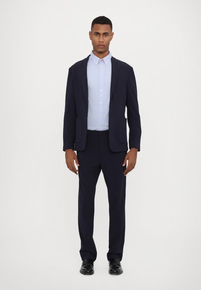 Costume marine avec un blazer ajusté et un pantalon assorti. Chemise bleu clair à boutons en dessous. Texture lisse et design classique. Chaussures de ville noires.