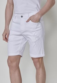 Shorts de denim blanco con un diseño desgastado, con dobladillos enrollados, bolsillos frontales y un cierre de botón en la cintura. Material ligero.