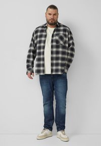 Kariertes Flanellhemd in Schwarz-Weiß, über einem weißen T-Shirt getragen. Kombiniert mit blauen Jeans und weißen Sneakers. Minimalistisches Design, lockere Passform.