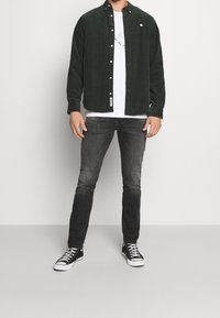 Grön kordflöjelskjorta med knäppning över en vit t-shirt, kombinerad med mörkgrå jeans och svarta låga sneakers.