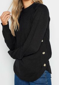 Svart stickad tröja med rund halslinning, ribbade ärmavslut och knappar på sidorna. Tyget har en mjuk textur och en avslappnad passform.