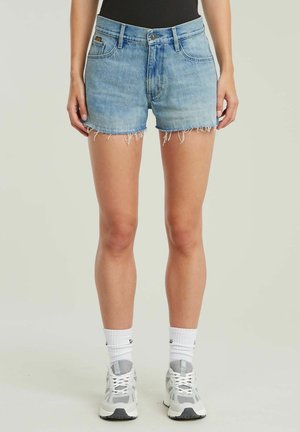Denimshorts in Hellblau mit ausgefransten Säumen, fünf Taschen und einem Knopfverschluss. Kombiniert mit weißen Sportsocken und Sneakers.