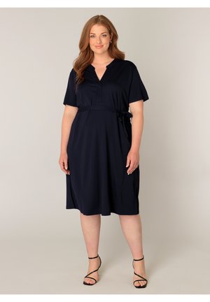 Vrouw met lang golvend haar, een marineblauwe jurk tot op de knie met korte mouwen en een zijstrik, gecombineerd met zwarte sandalen met bandjes.