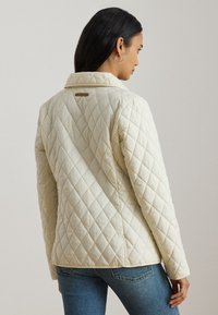 Femme aux cheveux foncés portant une veste matelassée crème et un jean bleu, vue de dos et légèrement de profil.