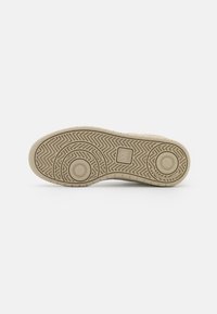 Semelle de chaussure beige avec des motifs de sculptures circulaires et en zigzag et un petit logo carré au centre sur un fond blanc uni.