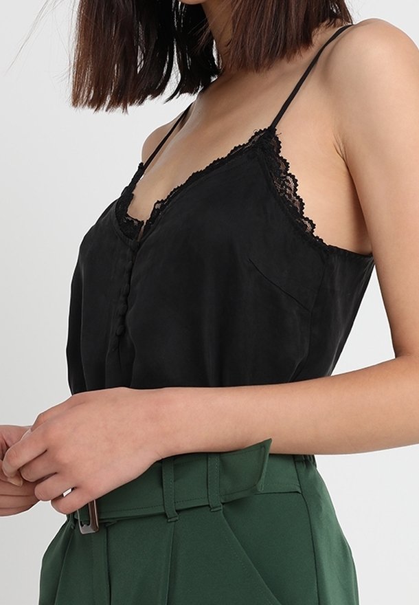 Camisole en soie noire avec dentelle et bretelles fines, présentant un devant à boutons et un détail de poche, associé à un pantalon taille haute vert.