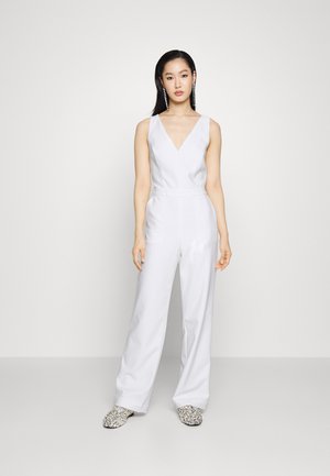 Witte mouwloze jumpsuit met een diepe V-hals, wijde broekspijpen en een aansluitende taille. Gedeeld met zilver versierde instappers. Gladde stoftextuur.