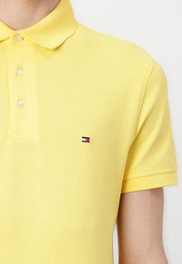 Gelbes Poloshirt aus strukturiertem Baumwollstoff, mit einem Drei-Knopf-Verschluss und einem kleinen gestickten Logo in Rot, Weiß und Blau auf der Brust.