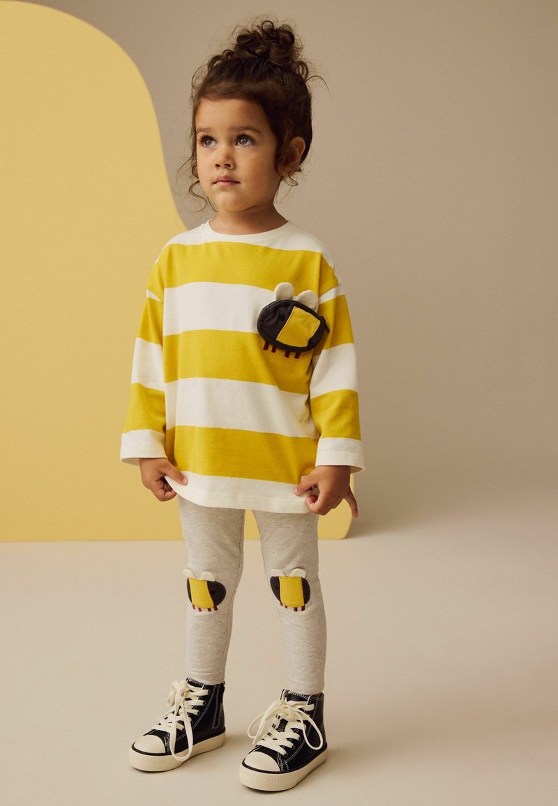 Langärmliges Shirt mit gelb-weißen Streifen und einer bienenförmigen Tasche, kombiniert mit grauen Leggings mit Bieneknieflicken und schwarzen Sneakers.
