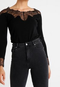 Haut noir avec décolleté en dentelle et accents de manches. Manches longues, design ajusté. Associé à un jean noir taille haute.