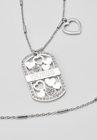 Collier chaîne en argent avec pendentif rectangulaire orné de plusieurs formes de cœur, certaines avec des strass, et "GUESS" gravé au centre.
