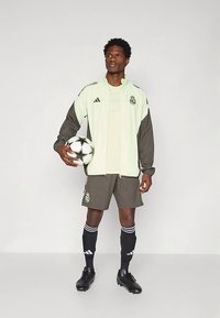 Ljusgrön och mörkgrå sportjacka med dragkedja, tillsammans med matchande shorts och svarta fotbollsskor. Håller i en fotboll.