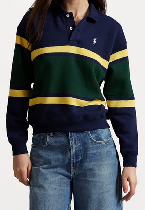 Mujer con un suéter tipo polo a rayas en azul marino, verde y amarillo, y vaqueros azules, de pie con las manos a los lados.