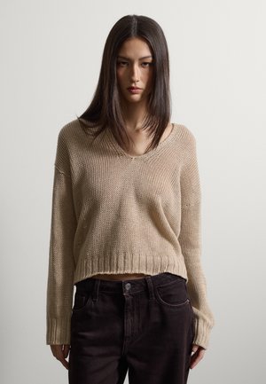 Vrouw met lang, stijl donker haar draagt een beige gebreide trui met V-hals en donkere broek, staand tegen een effen lichte achtergrond.