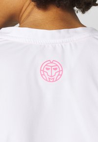 Bílá košile s růžovým kulatým logem připomínajícím masku, umístěná na zádech u výstřihu, s mírně patrnou texturou.