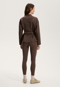 Brun idrottsjacka med bälte och matchande leggings. Slät, flexibel tyg; åtsittande design; rund hals; subtila sömdetaljer; låga fickor.