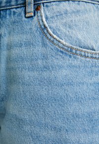 Tessuto di denim blu chiaro con una texture sottile, caratterizzato da una tasca con bordi sfilacciati e dettagli di cucitura color nocciola.