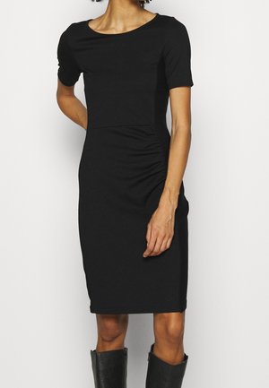 Kobieta w dopasowanej czarnej sukience sięgającej kolan, z krótkimi rękawami i subtelnymi marszczeniami po bokach, w połączeniu z czarnymi kozakami za kolano.