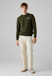 Olijfgroene sweatshirt met ronde halslijn en witte branding, gecombineerd met crèmekleurige cargobroek met zakken en een logo-opdruk. Casual sneakers.