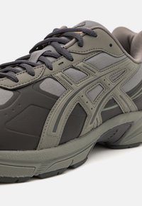 Chaussure de sport en gris foncé et olive, présentant une tige synthétique texturée, des accents en maille, une semelle épaisse et des lacets plats.