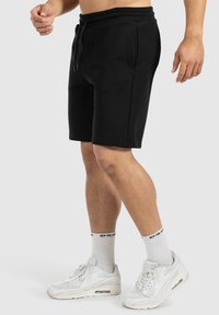 Schwarze Baumwollshorts mit einem Kordelzugbund und seitlichen Taschen, kombiniert mit weißen Sportschuhen und gerippten Socken.