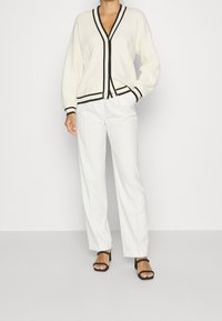 Bílý cardigan s černým lemem, výstřihem do V a zapínáním na knoflíky, kombinovaný s bílými širokými kalhotami a černými sandály na bloku.