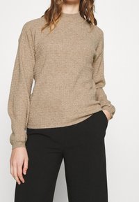 Beżowy sweter o teksturowanej powierzchni z wysokim dekoltem i długimi rękawami, w połączeniu z czarnymi spodniami. Charakteryzuje się luźnym krojem i ściągaczami przy mankietach.