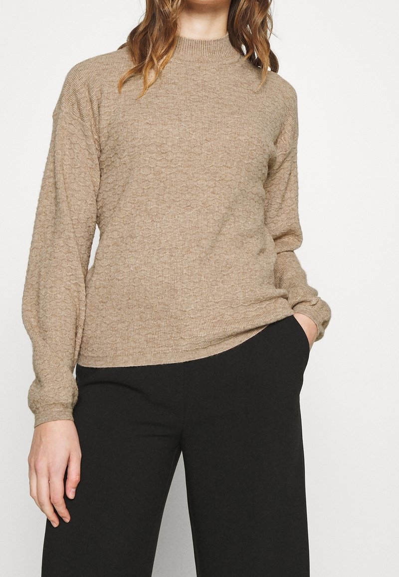 Beżowy sweter o teksturowanej powierzchni z wysokim dekoltem i długimi rękawami, w połączeniu z czarnymi spodniami. Charakteryzuje się luźnym krojem i ściągaczami przy mankietach.