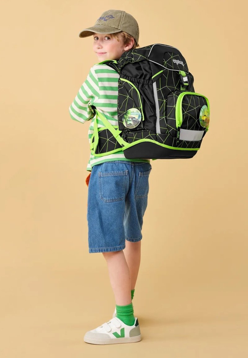 Black backpack with green geometric accents, multiple compartments, reflective strip, worn by a child in a striped shirt and denim shorts.