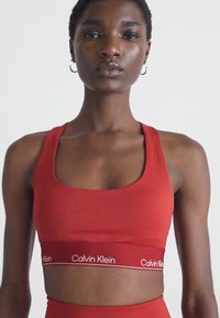 Femeie îmbrăcată într-un sutien sport roșu Calvin Klein cu bretele subțiri și bandă cu logo, stând pe un fundal simplu deschis.
