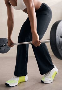 White athletic shoes with neon yellow accents, paired with black fitted pants. A barbell is held at waist level, with textured grip surfaces.