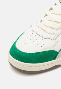 Białe skórzane trampki z zielonym zamszowym nakładką, perforacjami na palcach i kremowymi sznurowadłami. Charakterystyczna gumowa podeszwa zapewnia przyczepność.