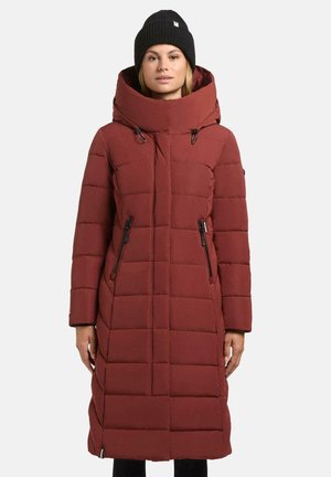 Vrouw in een lange bordeauxrode gewatteerde winterjas met een hoge kraag en capuchon, gecombineerd met een zwarte gebreide beanie.