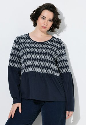 Mujer con cabello corto y rizado lleva una blusa azul marino con patrones blancos en zigzag y mangas largas, de pie frente a un fondo blanco.