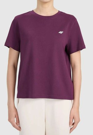 Kobieta ubrana w gładki fioletowy krótki rękaw z małym białym logo "4F" na lewej piersi, w zestawieniu z białymi spodniami.