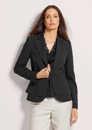 Frau mit langen braunen Haaren, die einen schwarzen Blazer über einem schwarzen Spitzenoberteil und cremefarbenen Hosen trägt, steht vor hellem Hintergrund.
