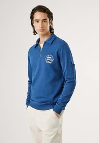 Mėlynas džemperis su užtrauktuku apykaklėje, su priekyje esančiu grafiniu spaudiniu. Jame yra gumuoti rankogaliai ir šoninis kišenės dizainas. Pagamintas iš medvilnės mišinio.