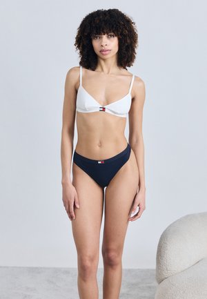 Woman with curly hair wearing a white bra and navy blue underwear with small logos, standing against a plain light gray background.