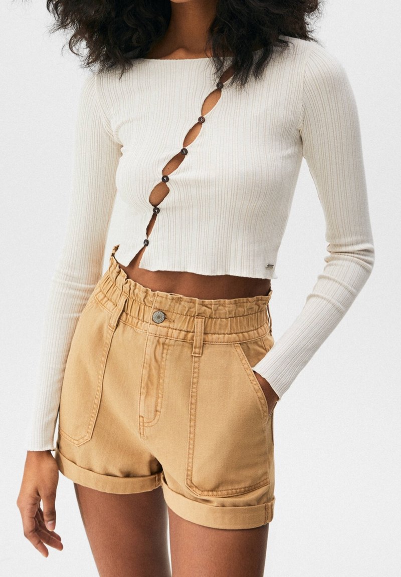 Femme portant un crop top blanc côtelé à manches longues avec des découpes asymétriques à boutons et un short beige taille haute style paperbag avec ourlets roulés.