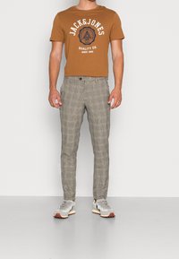 Braunes Grafik-T-Shirt mit weißem Text, kombiniert mit hellgrauen karierten Hosen und grauen Sneakers mit braunen Akzenten.