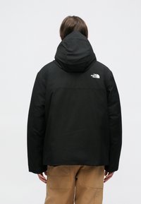 Zwarte hoodie met een gladde textuur en een wit logo op de bovenrug, gedragen over tan cargo pants. Eenvoudig, functioneel ontwerp.